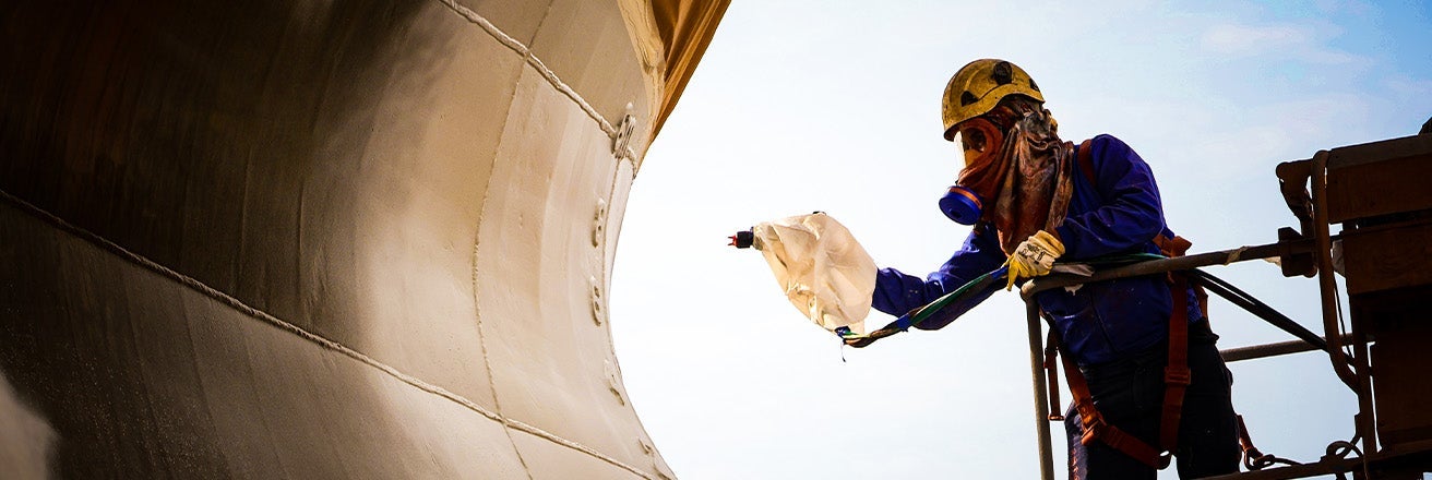 A person wearing a hard hat and blue uniform spraying paint on a ship 