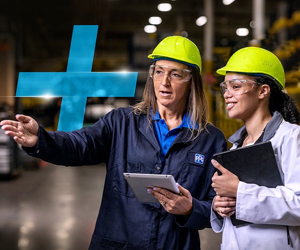 Two female workers in yellow hard hats and safety jackets in a manufacturing environment with a blue cross in the background
