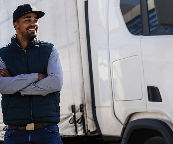 Truck driver with arms crossed standing confidently next to a white semi-truck in a sunny parking area