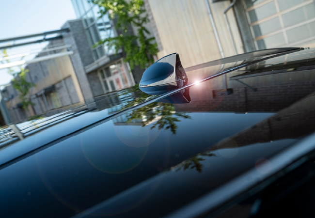 Close-up of a car roof finished in high-gloss black with a shark-fin antenna, reflecting nearby buildings and trees.