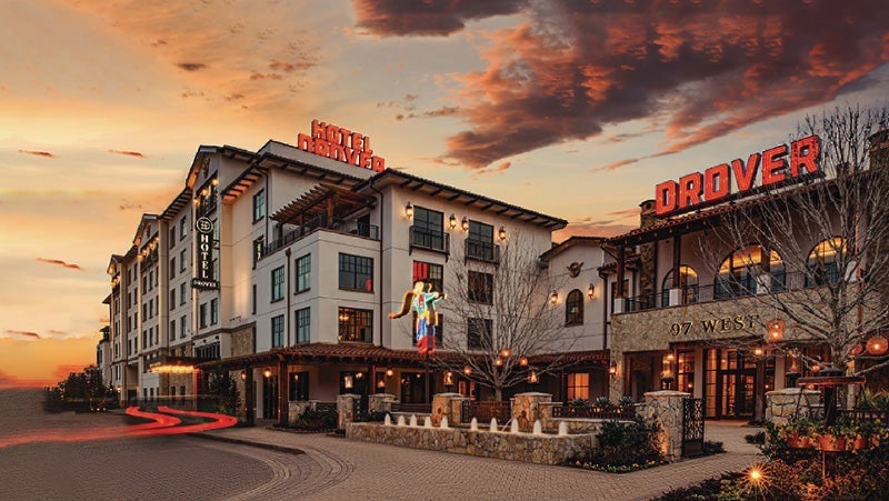 Twilight view of Hotel Drover with illuminated neon signs, cobblestone driveway, and vibrant sunset sky