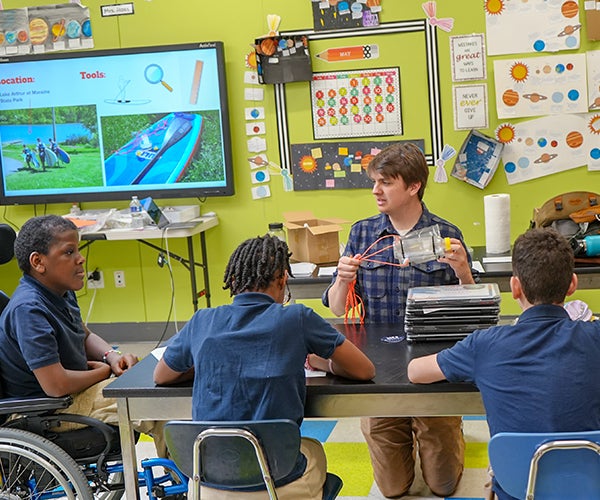 Male teacher demonstrating with apparatus in classroom to group of children