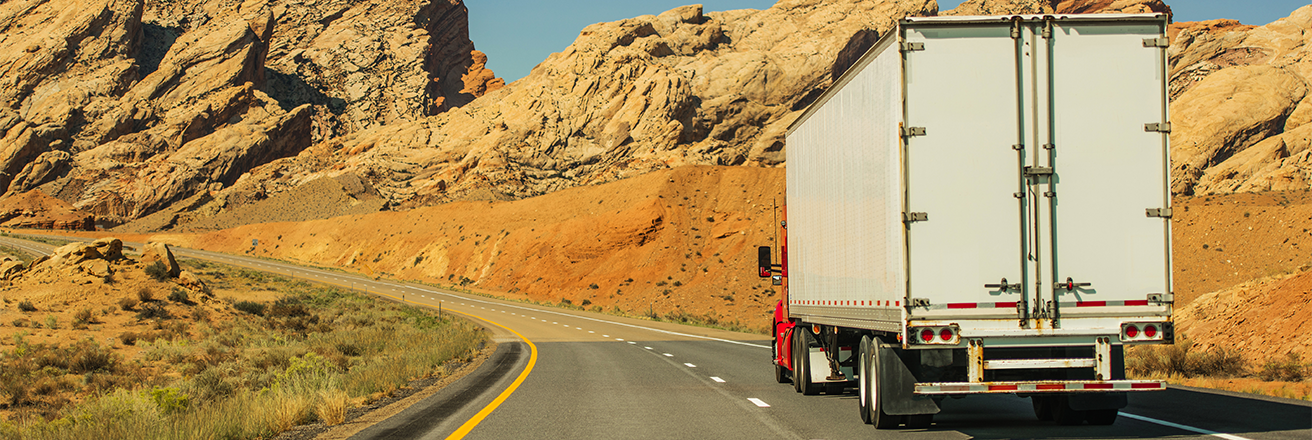 Transportation header image showing a white truck with a red cab driving up a sandy road surrounded by mountains