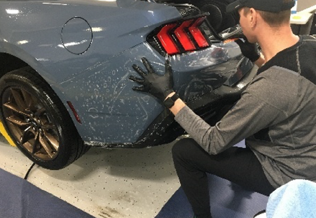 Technician applying protective film or refinishing material to the rear quarter panel of a gray car in an auto repair shop.
