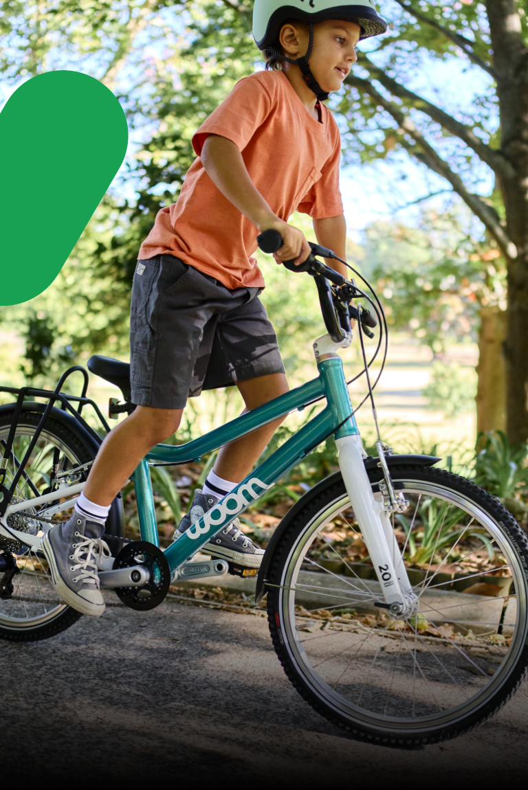 Un jeune garçon portant un casque READY vert roule en danseuse avec un woom GO 4 vert menthe sur une route bordée d’arbres.