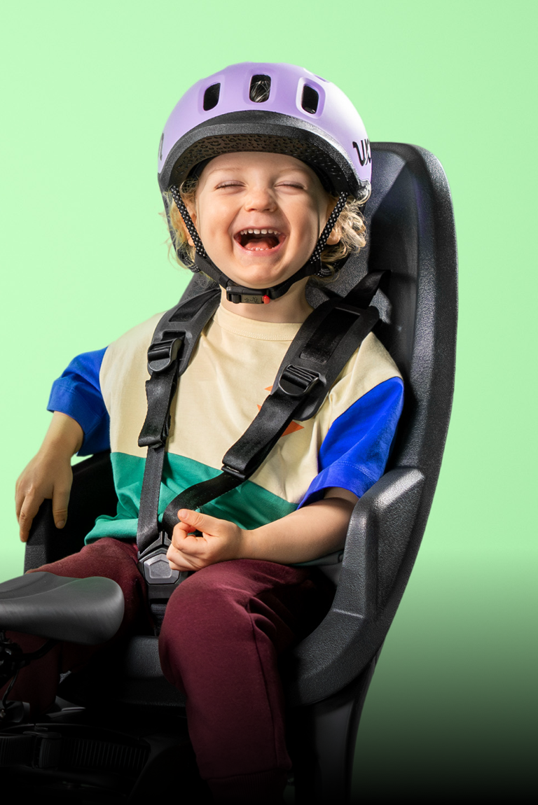 Photo d’un petit enfant heureux avec le casque READY de woom en violet, assis dans un siège vélo, arrière-plan vert