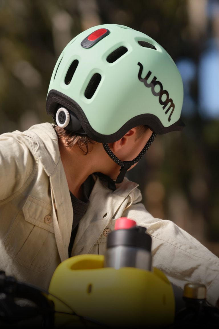 Vue de derrière d’un garçon portant le casque READY de woom de couleur vert menthe avec la lampe de casque rouge fixée dessus