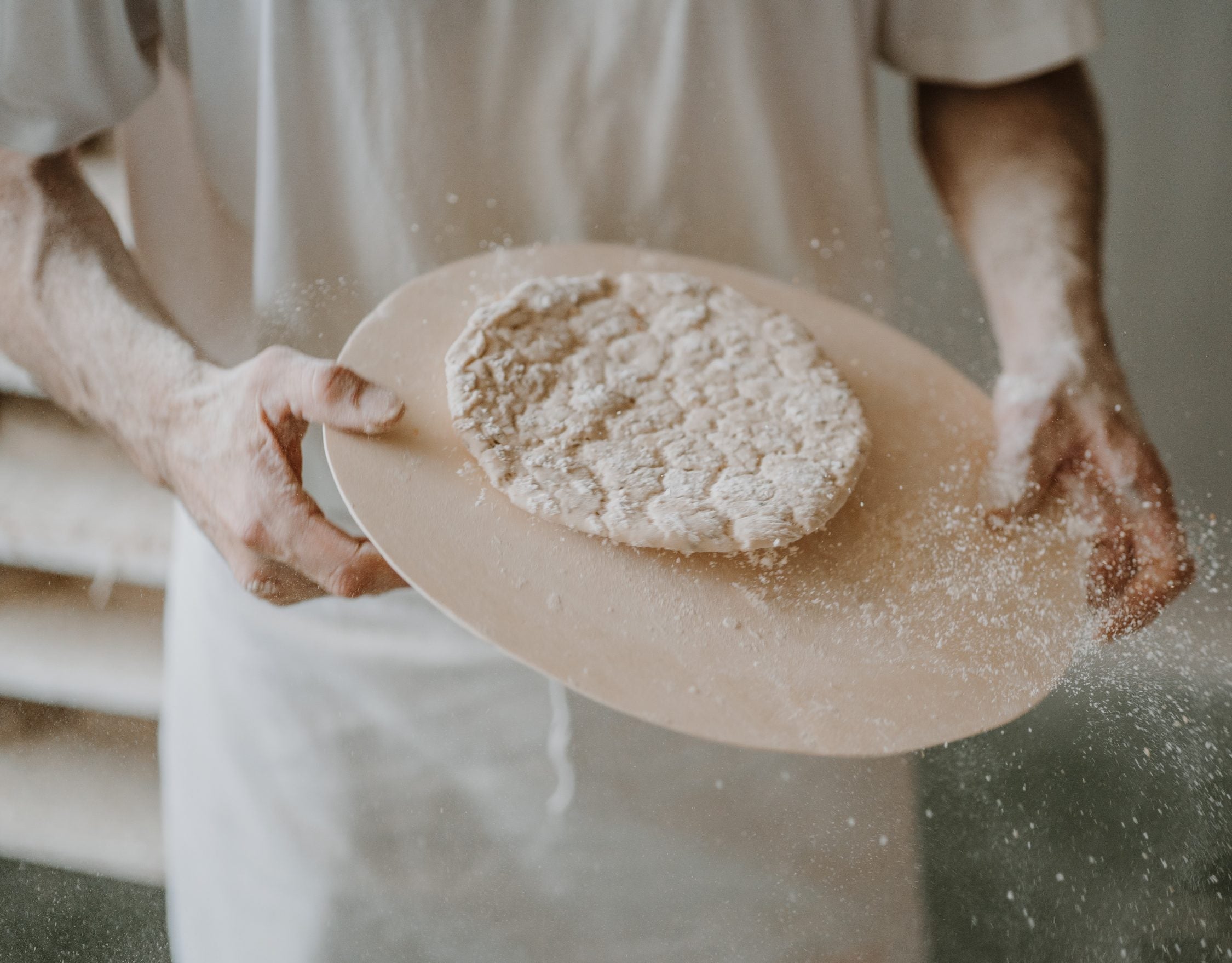 Bäcker schüttelt einen Schüttelbrotteig auf einer Holzschaufel, während Mehl in die Luft staubt.