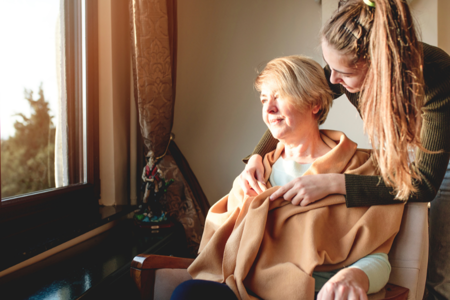 A woman sitting in a chair has a blanket draped over her shoulders by a younger woman. 