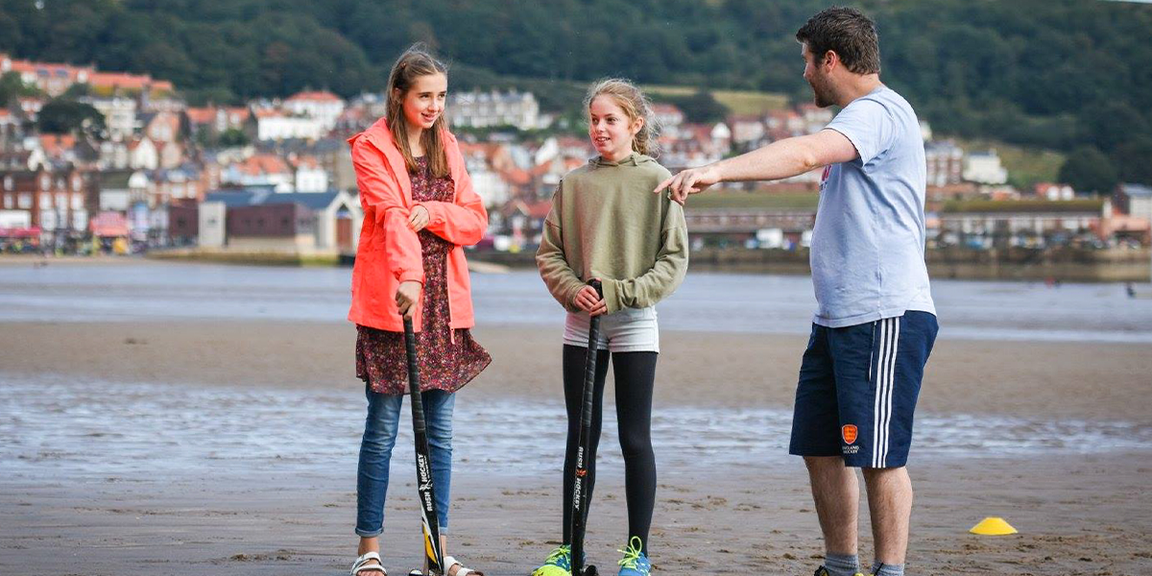 male player showing young players how to play hockey on the beach 
