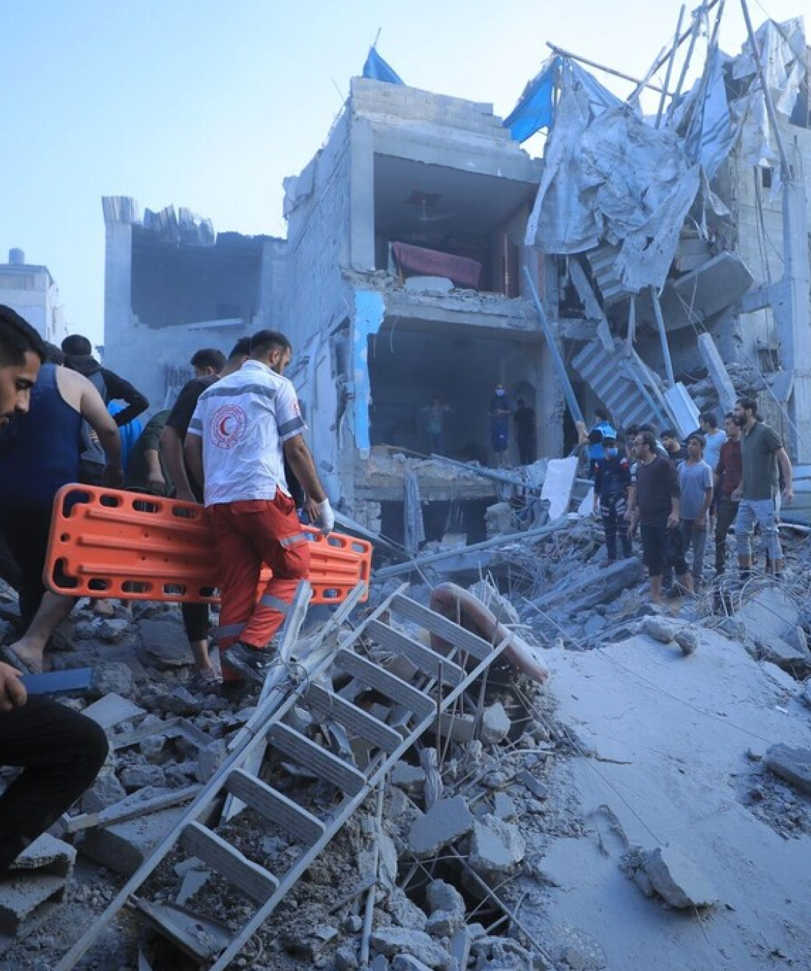 Palestinian Red Crescent Society workers carry a stretcher through rubble towards a house that has been bombed while a group of people watch on.