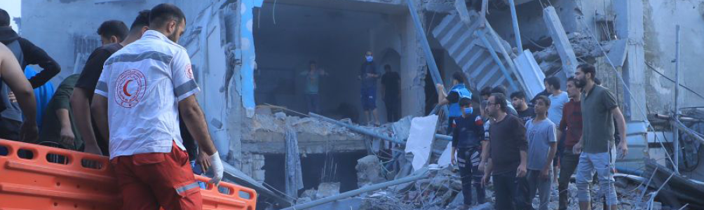 Palestinian Red Crescent Society workers carry a stretcher through rubble towards a house that has been bombed while a group of people watch on.