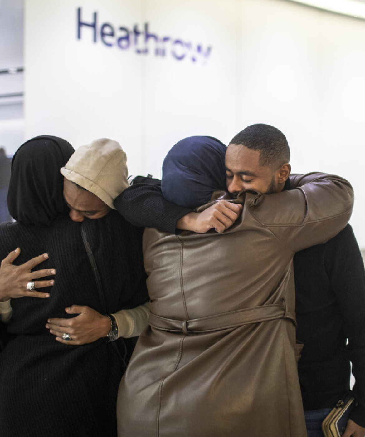 Two pairs of family members hug warmly in an airport terminal, leaning their heads together and smiling during a reunion.