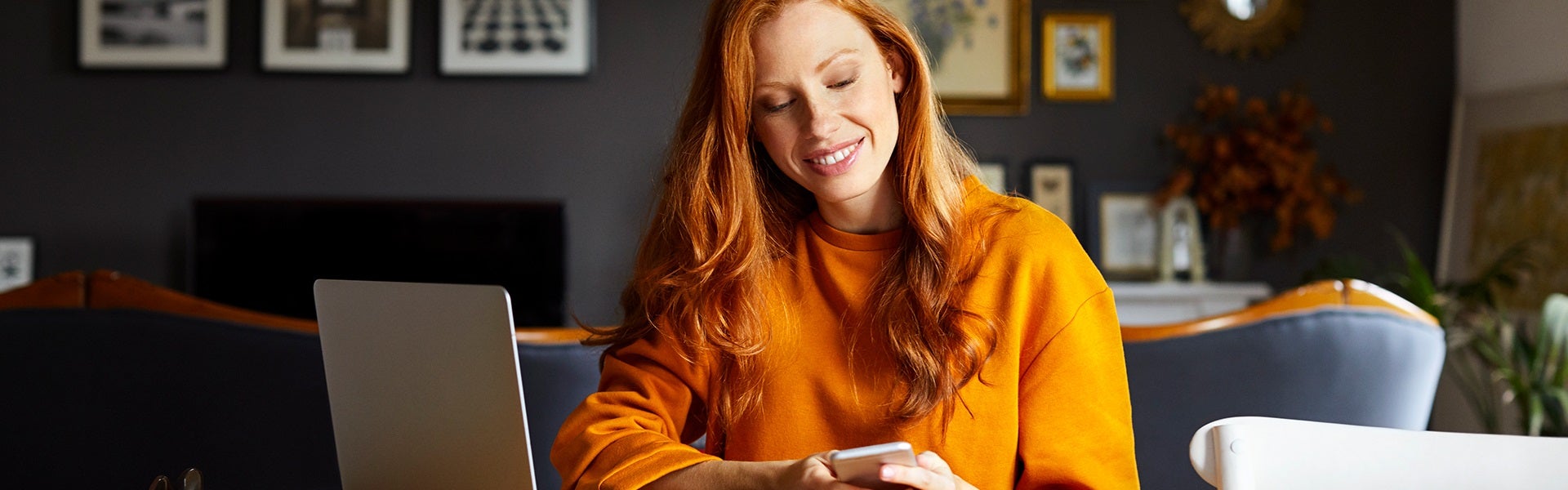 Vrouw in oranje trui kijkt op haar mobiel met laptop naast zich