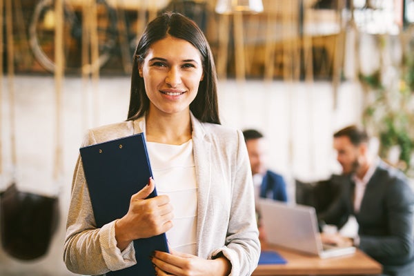 Jonge vrouw in beige jasje houdt blauw clipboard in armen terwijl op de achtergrond twee mannen praten rondom een laptop