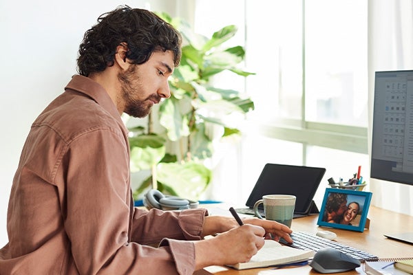 Man met bruin haar in lichtbruin overhemd schrijft aan een bureau met daarop een kop koffie, laptop en fotolijst