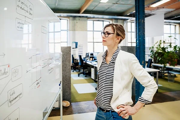 Vrouw met blond haar en bril in zwart-wit gestreepte blouse staat met handen in haar zij voor een whiteboard