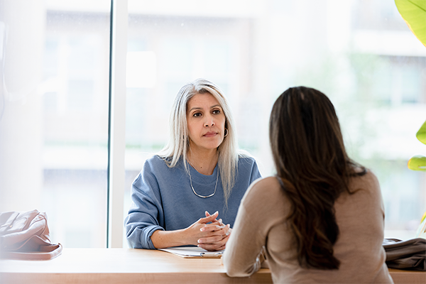 Vrouw met wit haar in blauwe blouse zit met handen gevouwen achter bureau tegenover een vrouw met bruin haar