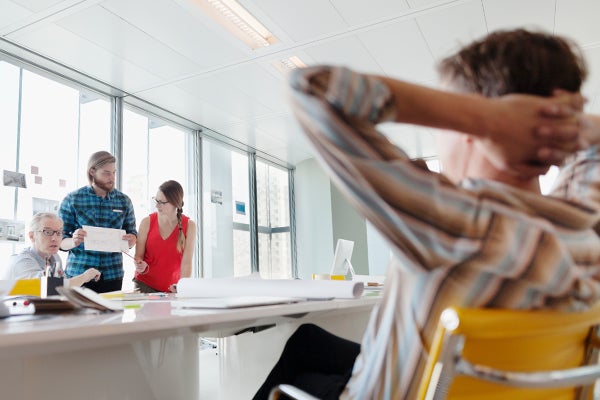 Man in bruin-blauw gestreept hemd leunt achterover en kijkt naar groepje van drie personen dat achter een bureau staat te overleggen
