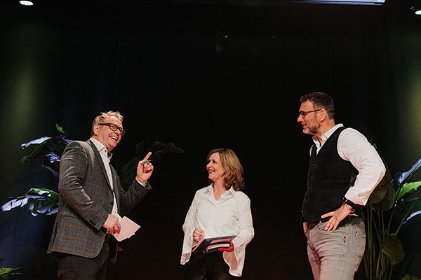 Drie sprekers op een podium; links een man die een handgebaar maakt, daarnaast een vrouw en man die vriendelijk lachend naar hem luisteren