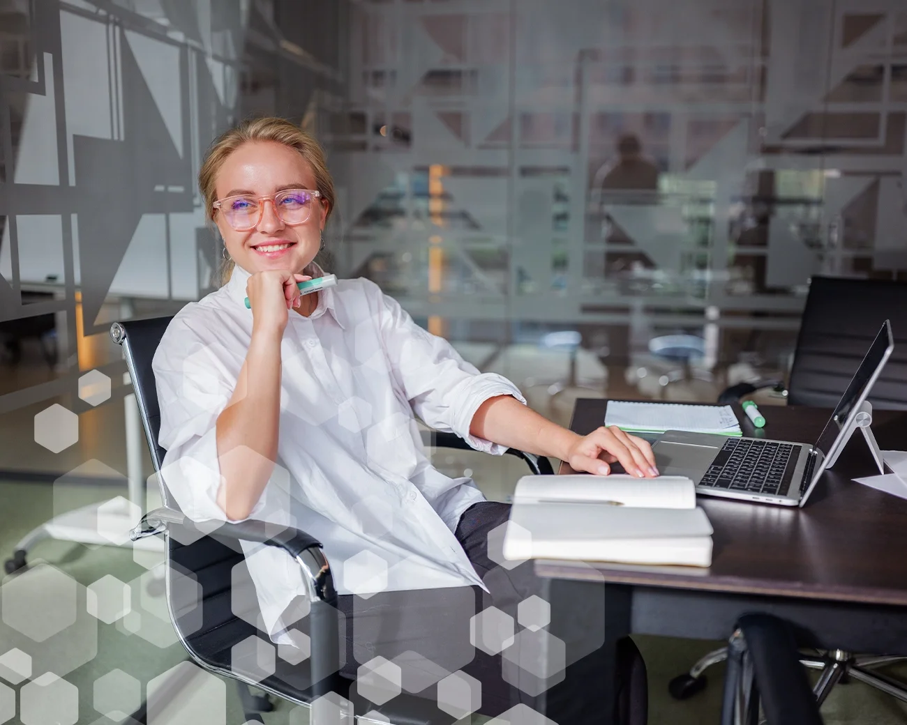 Jonge vrouw met bril zit achter bureau met boek en laptop