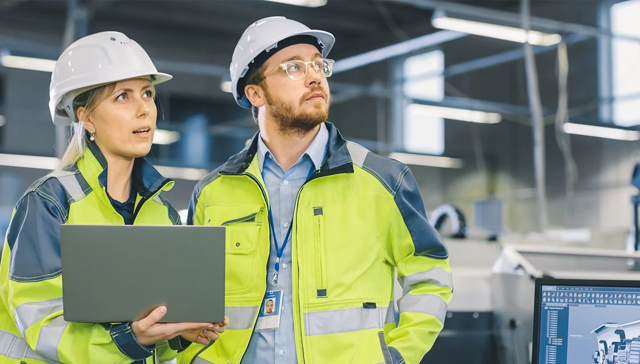 Man en vrouw met bouwhelm en laptop in fabriekomgeving