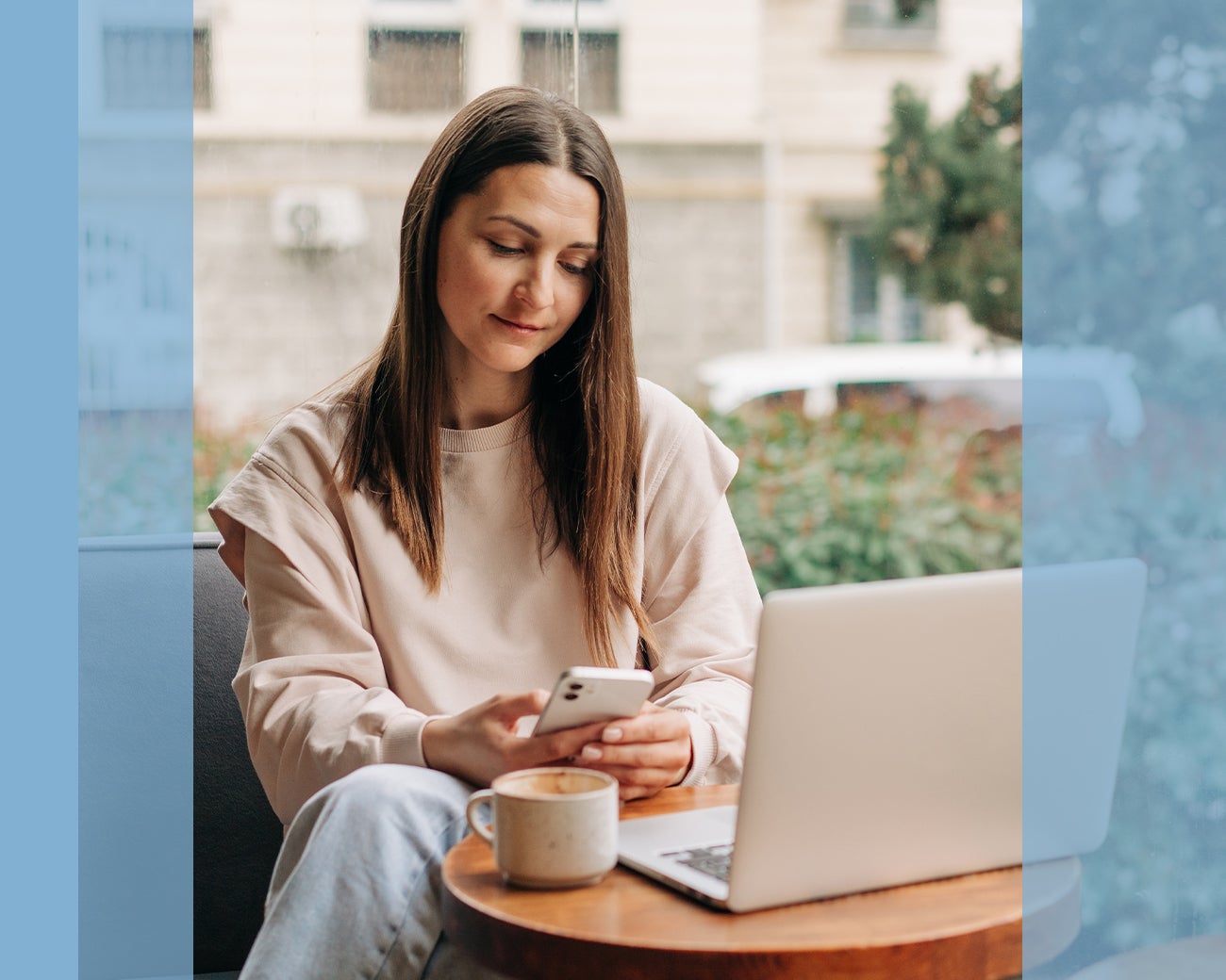 Jonge vrouw zit aan tafel met koffie en laptop, kijkt op telefoon