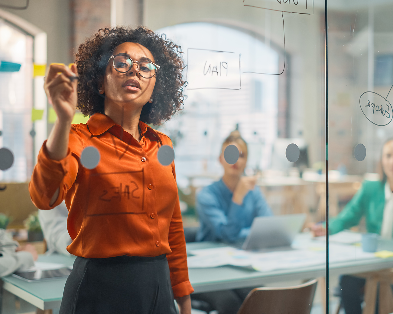 Vrouw met krullend haar en bril en oranje blouse schrijft op een raam