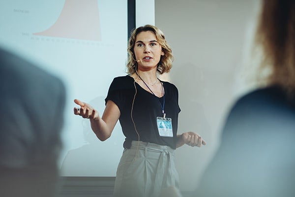 Vrouw met kort blond haar in zwart shirt staat voor een scherm en spreekt met microfoon een publiek toe