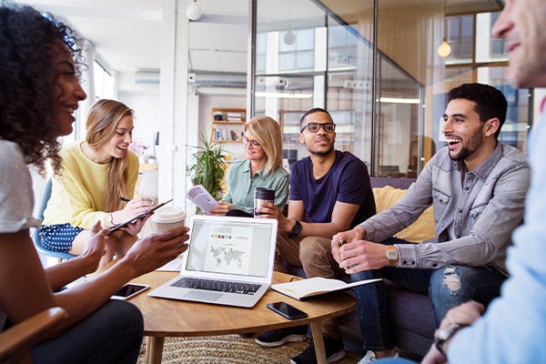 Groep mensen lachend rondom een laptop in een ruimte met houten tafel en glazen wanden