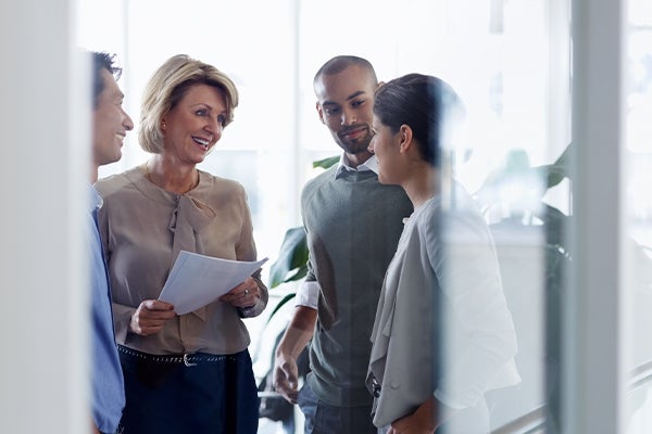 Vier mensen in een lichte ruimte. Middelbare vrouw in grijze blouse met stapel papieren in haar hand praat vriendelijk lachend tegen een jonge vrouw met bruin haar.