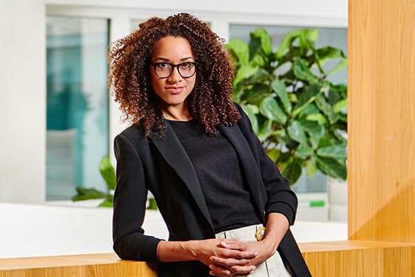 Vrouw met bruin haar en bril leunt achterover op een vensterbank met plant op de achtergrond