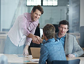 Man in roze overhemd staat over tafel gebogen en schudt hand van persoon met kort bruin haar in blauw jasje