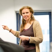 Vrouw met beige jasje en bruin shirt staat en wijst naar links