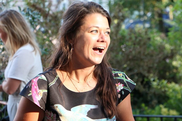 Vrouw met bruin haar en blouse met kleurrijk vogelmotief staat buiten in de zon en lacht