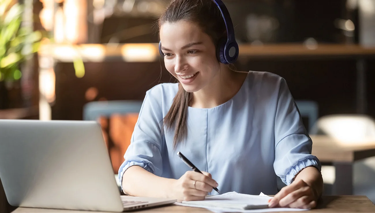 Vrouw met koptelefoon maakt notities terwijl ze naar haar laptop kijkt