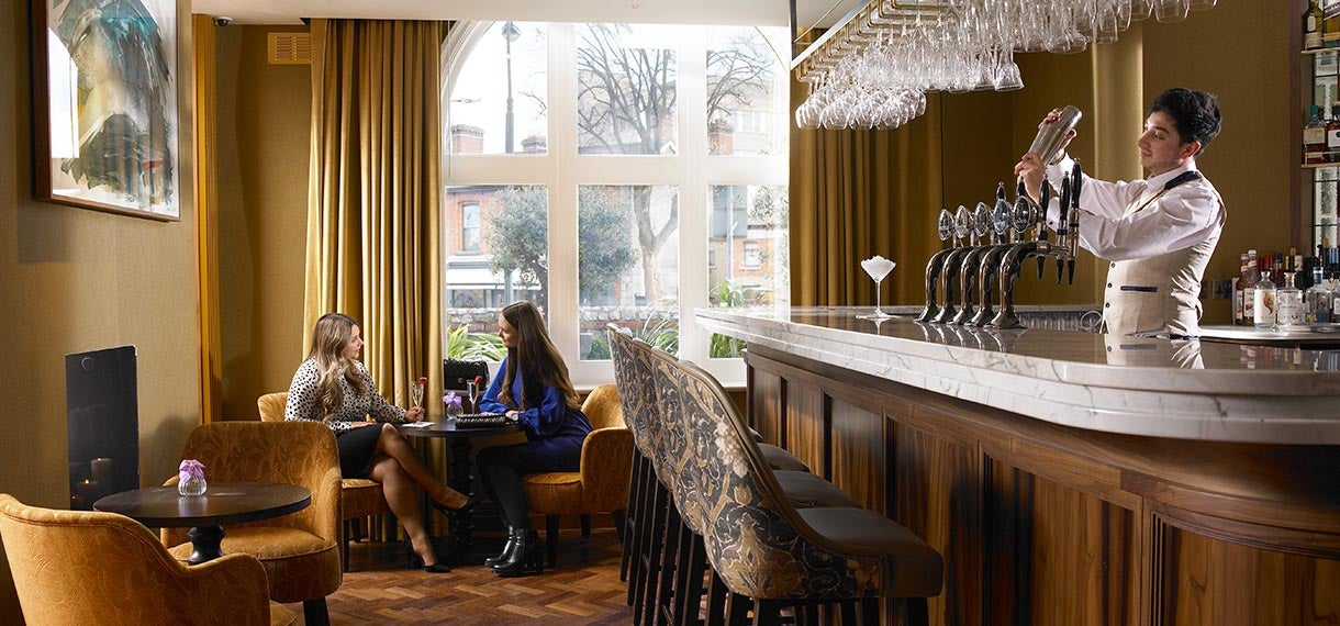 Bartender mixing a drink while two women sit and talk at a table in a stylish bar.