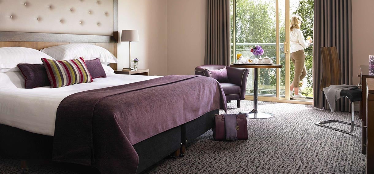 Elegant hotel room with a purple theme, large bed, table, and window with a person enjoying the view.