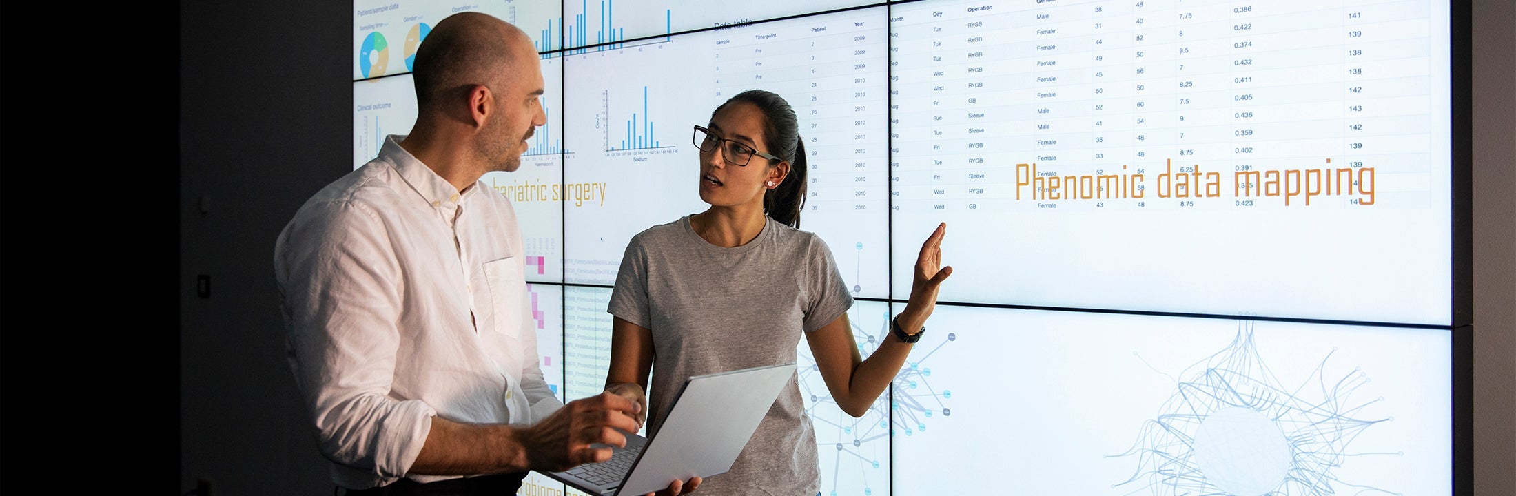 Two colleagues talk to each other in front of a screen that displays data