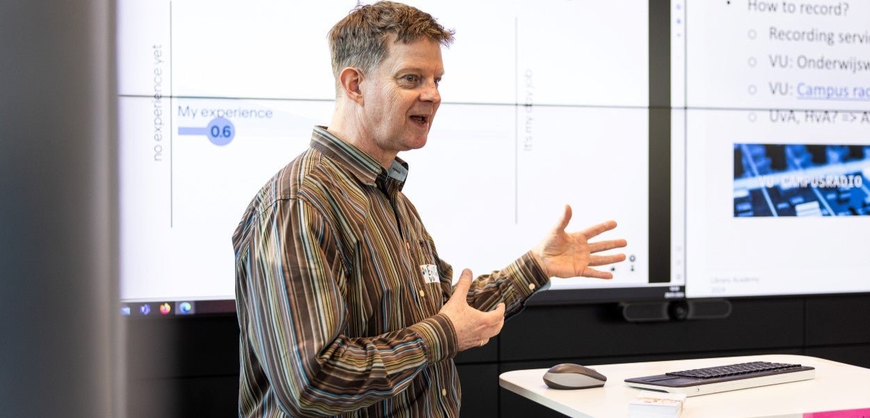 Person in front of a presentation screen with texts and graphics, gesturing with hands and speaking.