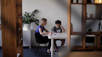 Blonde woman with blue shirt and pen in hand sits at white table with a boy with black curls