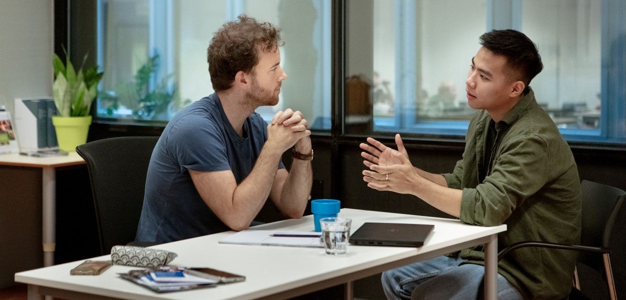 Two people are sitting across from each other at a table; one is talking and the other is listening