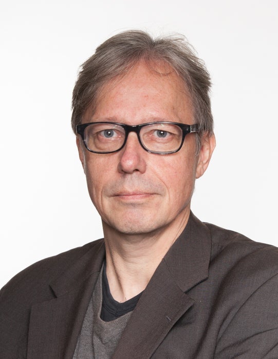Portrait photograph of a man with gray hair and dark glasses, dressed in a dark beige jacket and gray shirt