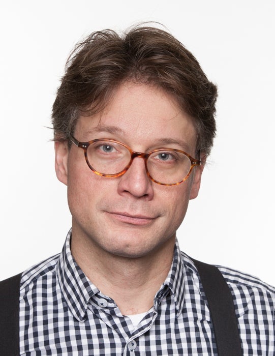 Portrait photograph of a man with brown hair and light brown glasses dressed in a black and white plaid shirt and black suspenders