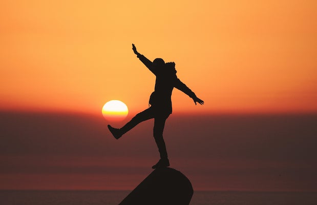 Person stands balancing on top of rock against vivid sunset backdrop