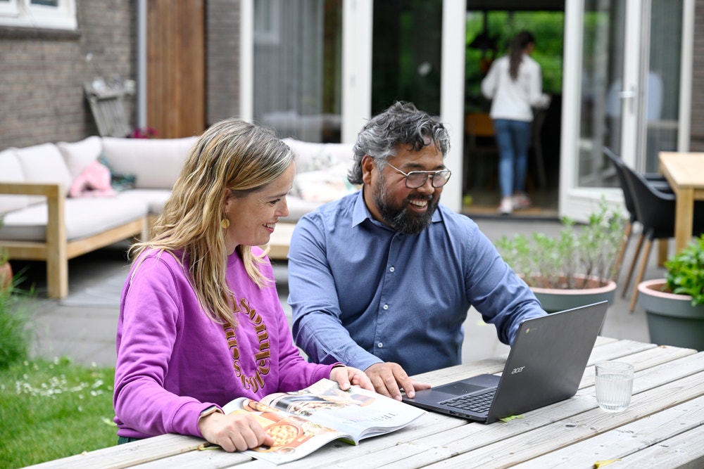 Man en vrouw zitten buiten aan een tafel en kijken lachend naar een laptop.