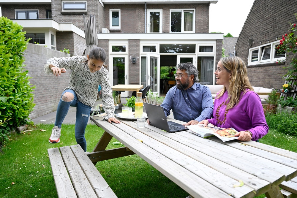 Man en vrouw zitten buiten aan een tafel en een meisje springt op de bank.