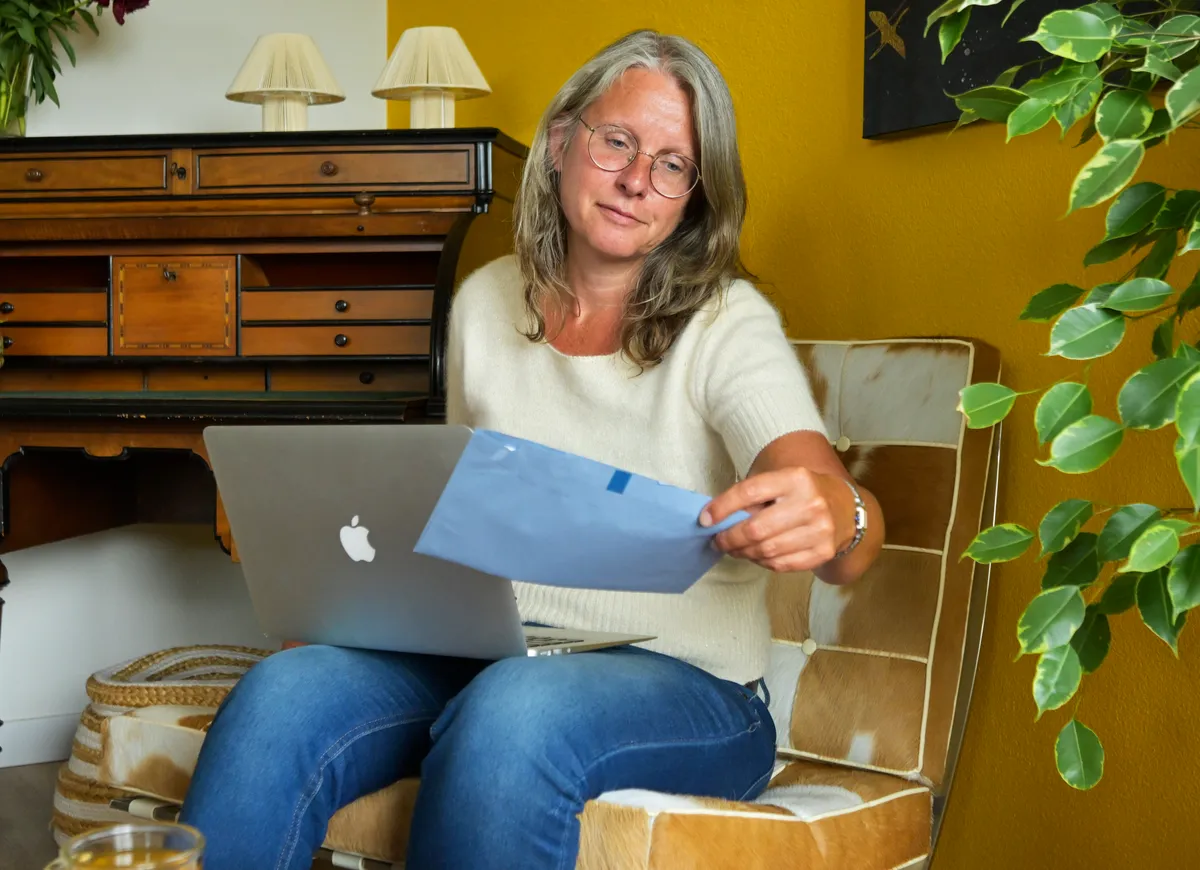 Vrouw op stoel in woonkamer met laptop op schoot bekijkt blauwe envelop van de Belastingdienst