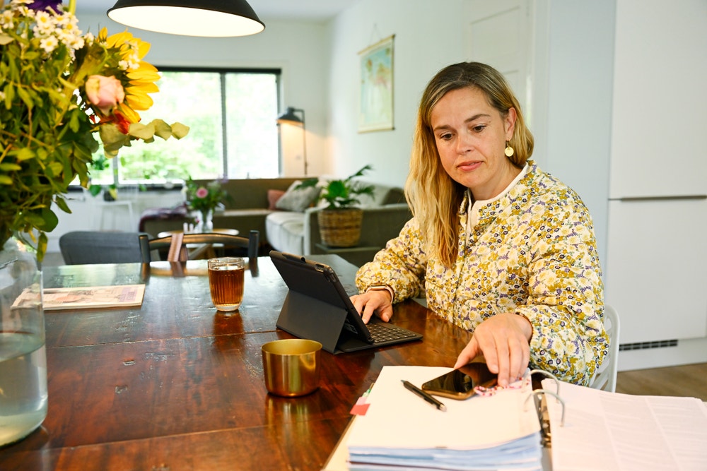 Vrouw zit aan tafel met tablet en een map. Ze pakt haar telefoon.