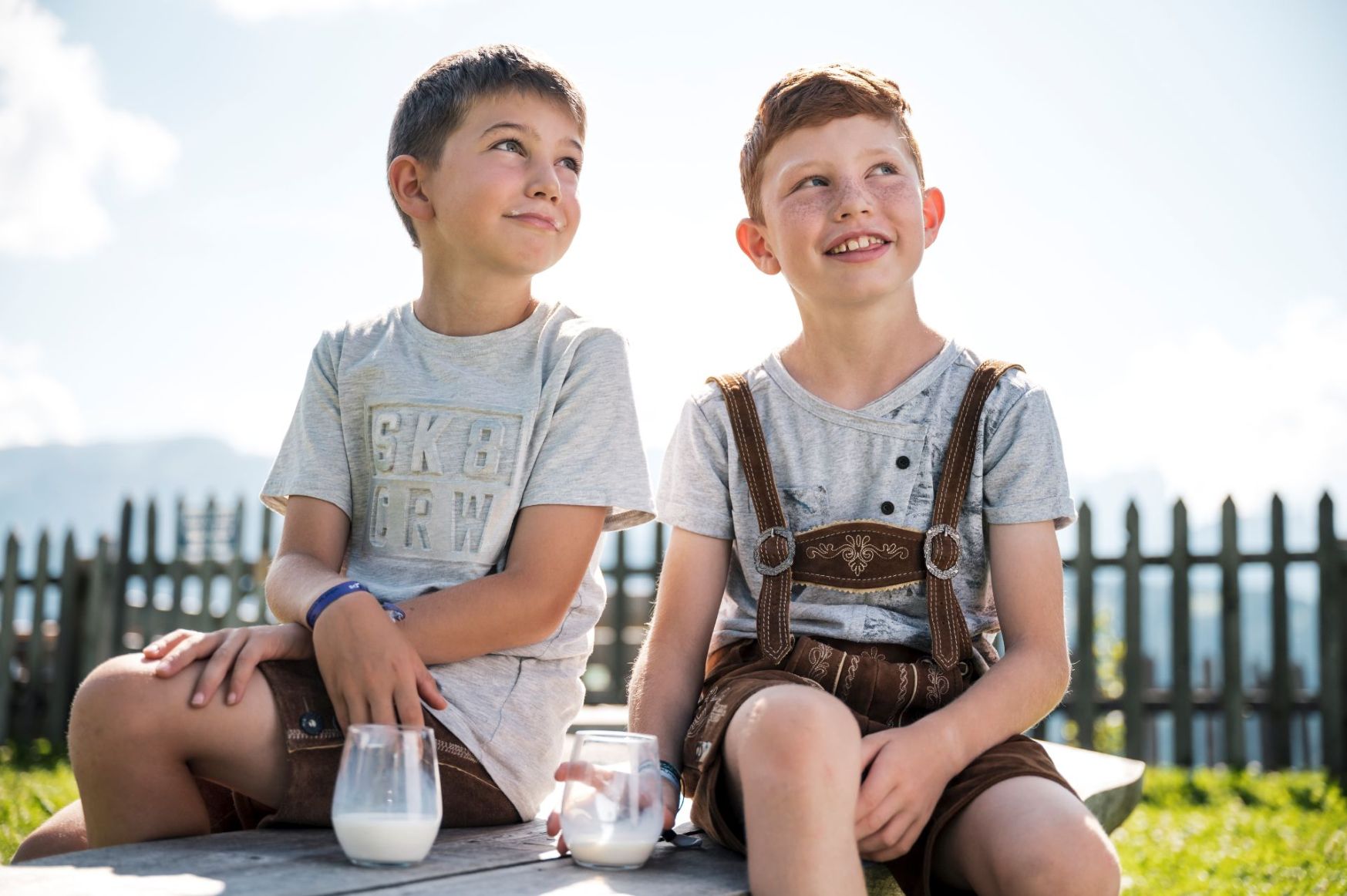 Zwei lächelnde Kinder sitzen auf einer Holzbank, jedes mit einem Glas Milch.